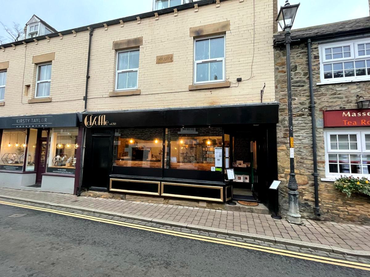 Coffuffle's shopfront on Middle Street, Corbridge: a black frontage with gold lettering and lit windows, in a pale stone terrace